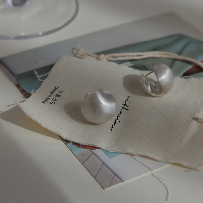 Close-up of brushed dome-shaped silver earrings with a matte finish from STUD, displayed on a white cloth backdrop.