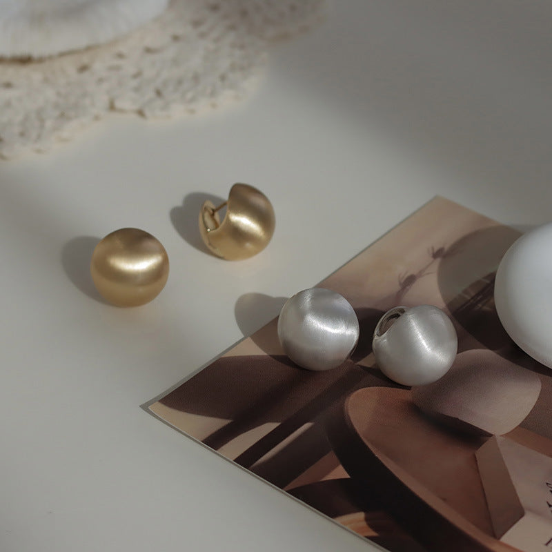 Close-up of brushed dome-shaped silver earrings with a matte finish from STUD, displayed on a white cloth backdrop.