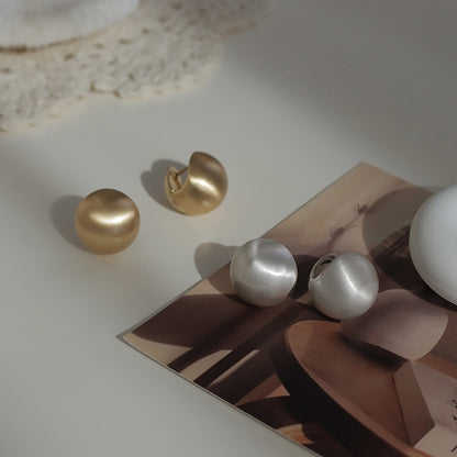 Close-up of brushed dome-shaped silver earrings with a matte finish from STUD, displayed on a white cloth backdrop.