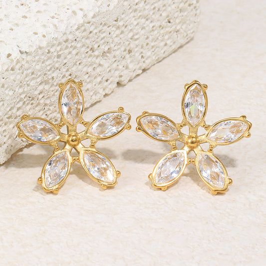 Gold flower-shaped earrings with clear gemstones on a textured beige surface